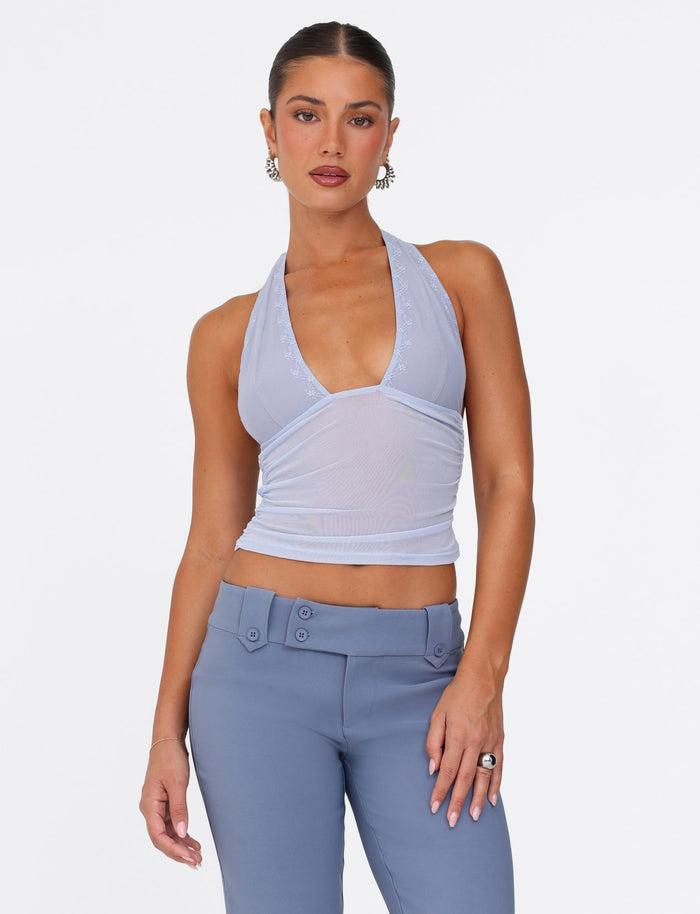A person poses against a plain white background, dressed in a light blue steel-coloured Bresily top by Tigermist and high-waisted blue pants. Their hair is tied back, and they are accessorized with hoop earrings and a ring.