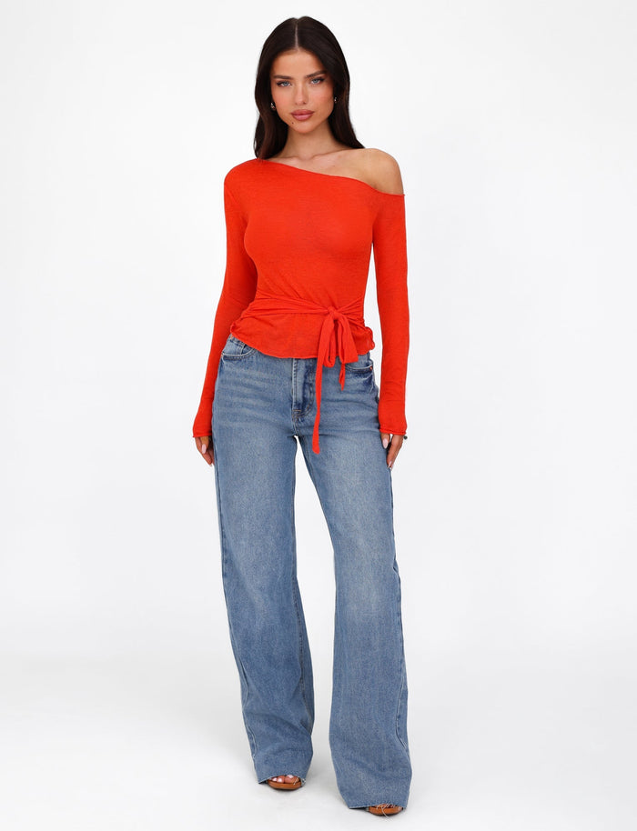 A woman stands against a plain white background, wearing the BRYNA JUMPER in red by TIGERMIST, an off-the-shoulder long-sleeved top with a waist tie, paired with blue high-waisted wide-leg jeans. The fitted silhouette of her outfit highlights her long dark hair as she looks directly at the camera with a neutral expression.