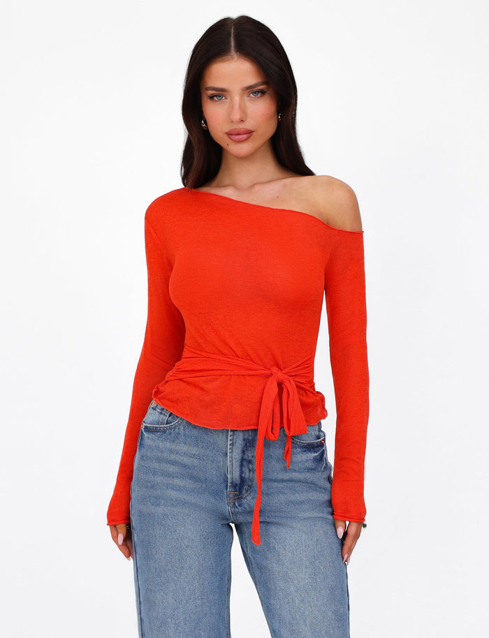 A woman with dark hair is wearing the bright red "BRYNA JUMPER" from TIGERMIST, an off-shoulder, long-sleeve top featuring a tied waist detail that flaunts a fitted silhouette, paired with blue jeans. She stands against a plain white background.