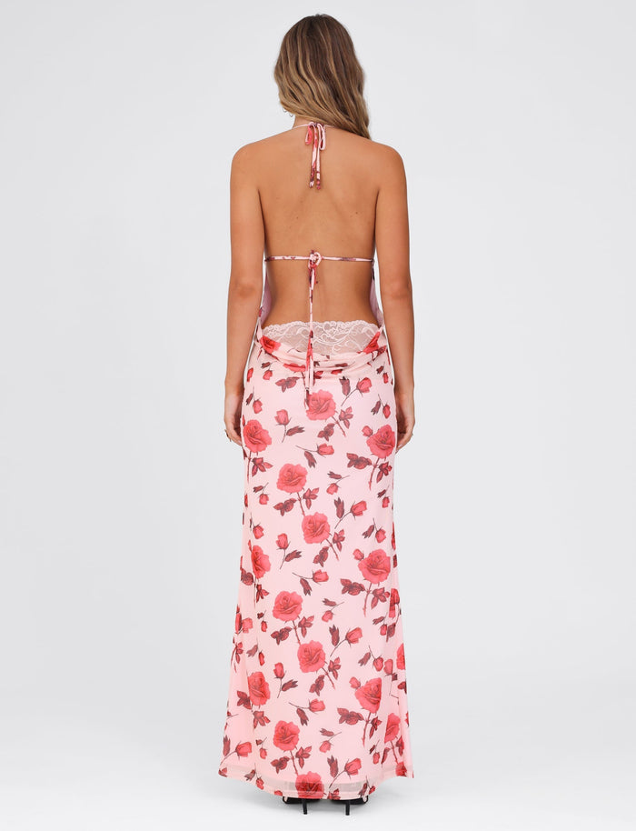 A woman with long hair stands facing away, wearing the TIGERMIST BRYSTOL DRESS - RED : FLORAL. This maxi-length dress showcases a halter neckline, a low back, and features a floral pattern in shades of pink and red with a flowing, full-length skirt.