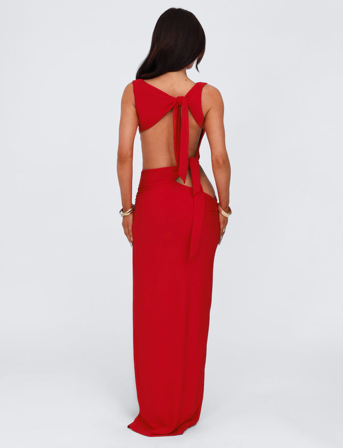A woman with long dark hair stands with her back to the camera, showcasing the TIGERMIST CASPIAN DRESS in red. This floor-length gown features an open back with two tied knots and minimal detailing, complemented by a high split that adds elegance. She accessorizes with a bracelet on one wrist, set against a plain white background.
