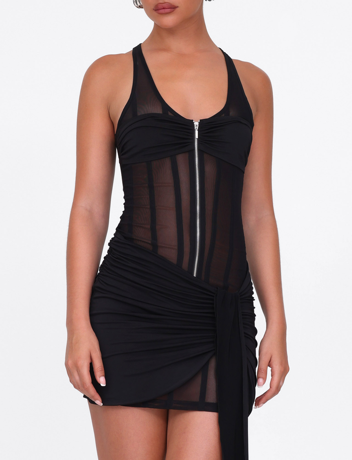 A person stands against a plain white background wearing the TIGERMIST CATERINA MINI DRESS in black—a sleeveless, sheer mesh mini dress with a zipper front, ruched skirt detailing, and slinky jersey lining.