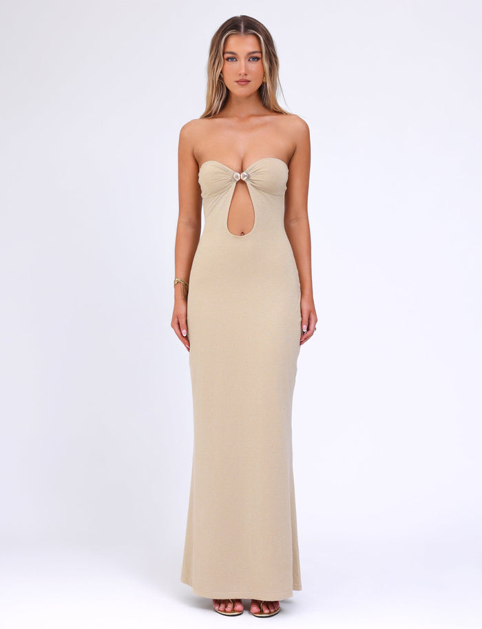 A woman stands posing in the CECILY MAXI DRESS - GOLD by TIGERMIST, a floor-length, fitted beige gown featuring a circular ring at the bust and a keyhole cutout below. The plain white background accentuates the dress's elegant design and its luxurious gold colourway.