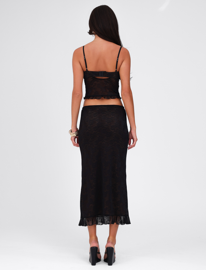 A woman with long brown hair faces away, wearing the TIGERMIST CHARLEIGH SKIRT - BLACK: LACE with a matching black lace cami top and high heels, set against a plain white background.