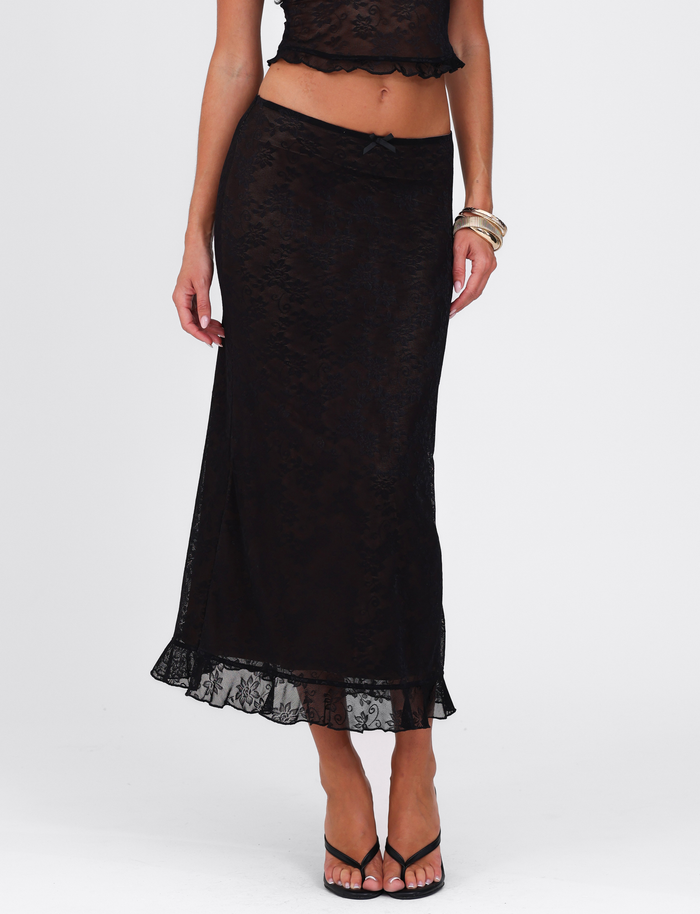 A woman wears the TIGERMIST CHARLEIGH SKIRT - BLACK in lace with a matching crop top, black sandals, and bangles, standing against a plain white background.