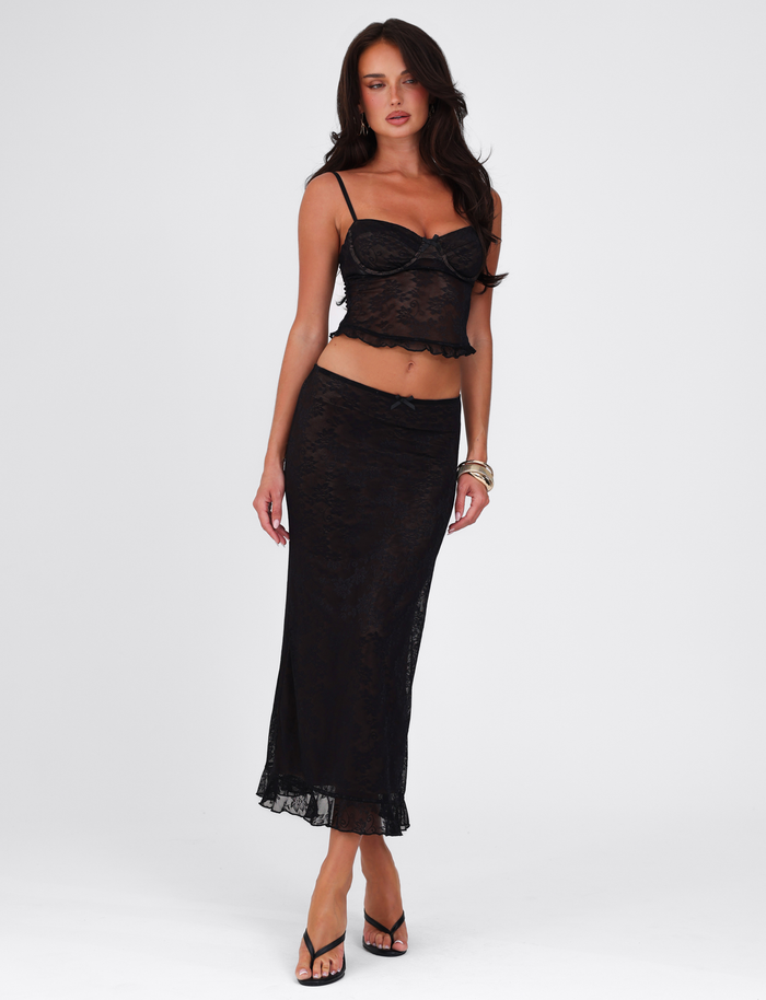A woman poses against a plain background, wearing the TIGERMIST CHARLEIGH SKIRT - BLACK: LACE with a matching black lace camisole, paired with black heeled sandals, long dark hair, and gold bracelets.