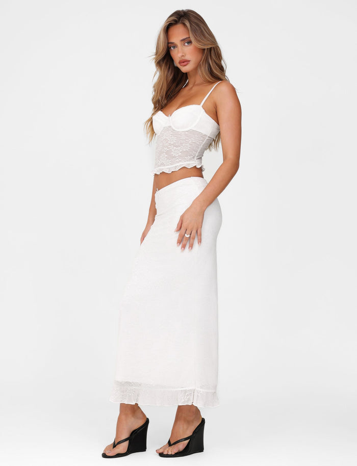 A person wearing the TIGERMIST CHARLEIGH SKIRT - WHITE : LACE and a white lace crop top stands against a simple backdrop. The outfit, with its delicate low-rise waist design, is elegantly paired with black wedge sandals. Their long hair flows down gracefully.