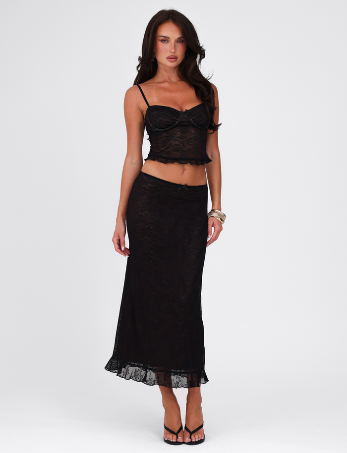 A woman models the TIGERMIST CHARLEIGH TOP - BLACK : LACE, a black lace crop top with adjustable straps, paired with a matching long lace skirt. She stands against a white background with loose wavy hair, bracelets, and black sandals.