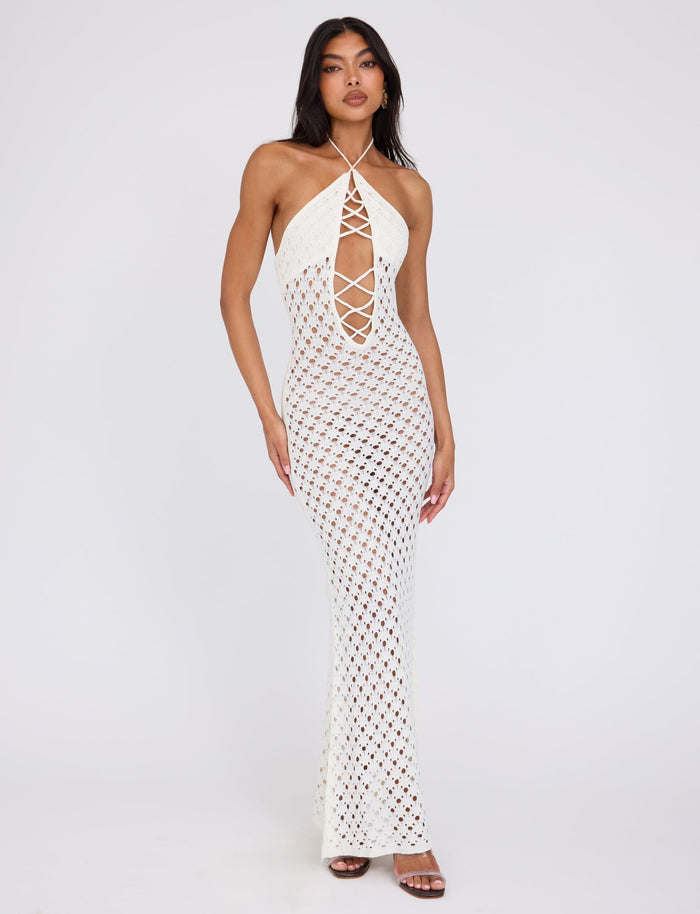 A woman stands against a plain white background, wearing the CIARAN DRESS - WHITE by TIGERMIST. The dress features a long, sleeveless halter-neck design with a lattice pattern and a deep crisscrossed cutout in the front. She has long dark hair and pairs it with brown high-heeled shoes.