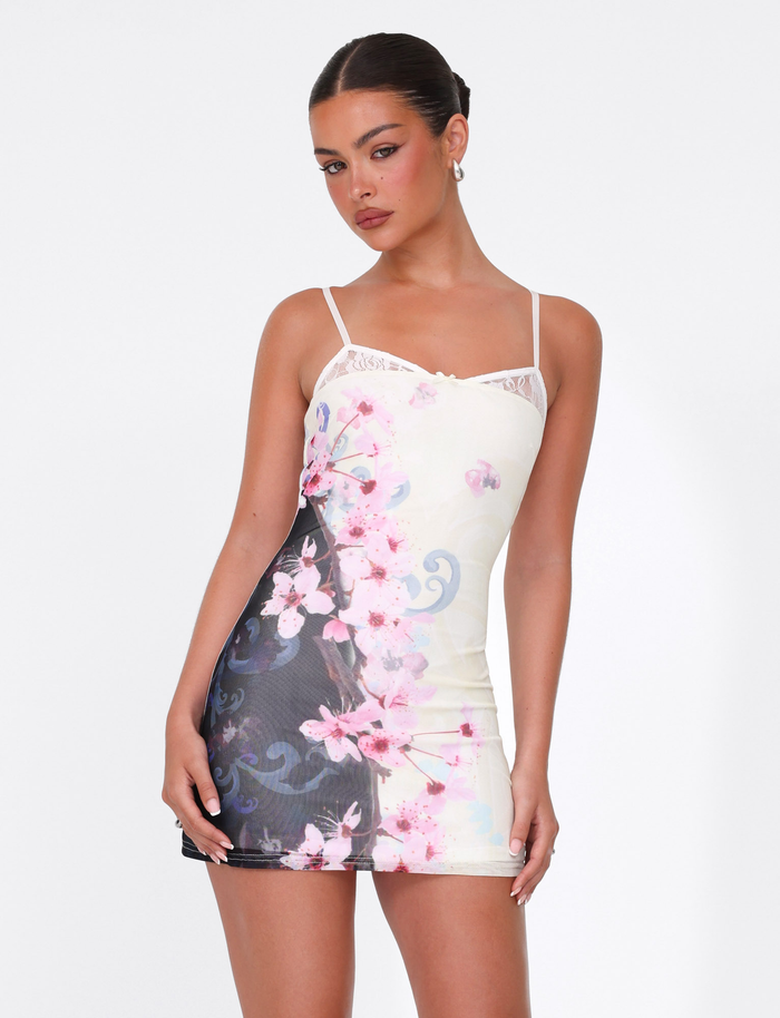 A woman with dark hair in a sleek bun models the TIGERMIST COLSON MESH MINI DRESS in multi cherry blossom floral, with adjustable straps and pink and blue designs, standing out against a plain white background as she looks to the side.