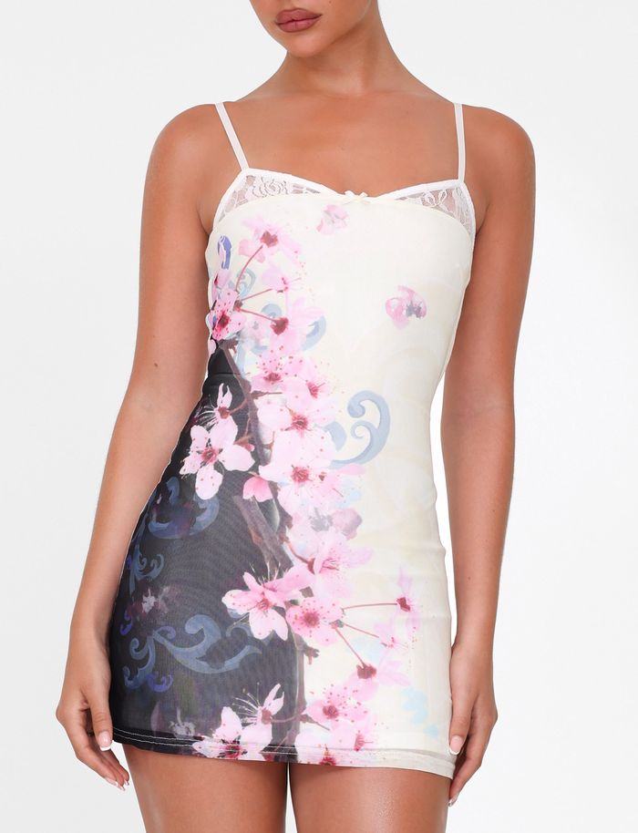 A woman wears the TIGERMIST COLSON MESH MINI DRESS in Multi: Floral Cherry Blossom, a sleeveless, fitted mini dress with adjustable straps and pink floral prints on black and white, standing against a plain white backdrop.