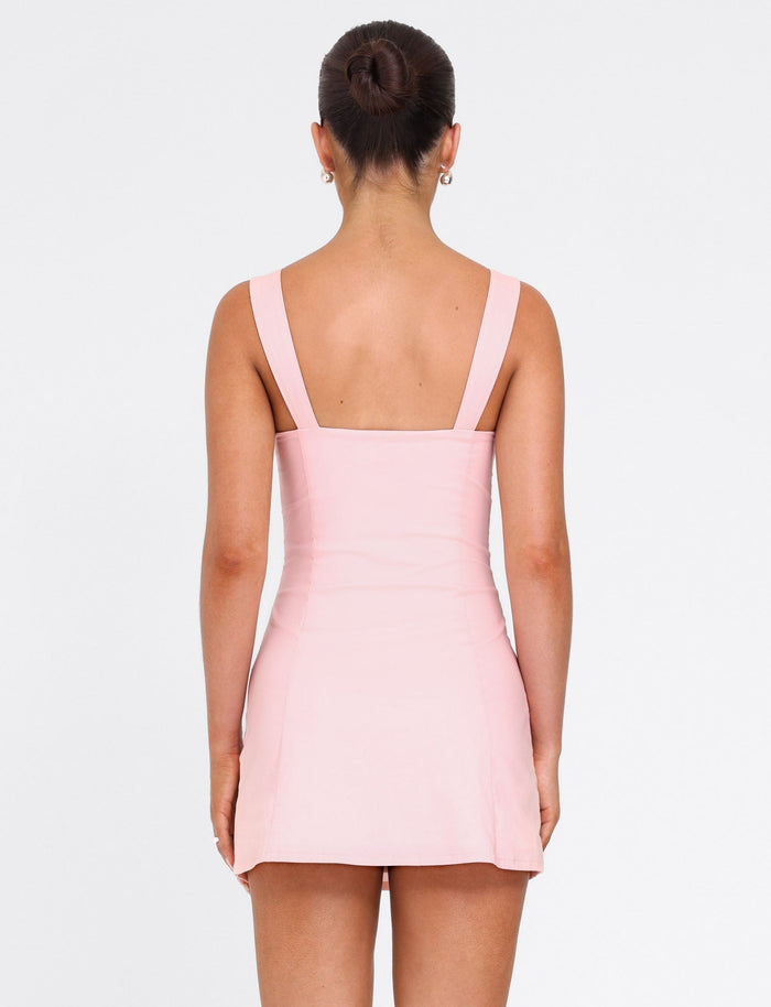 A woman with dark hair in a bun is seen from behind, wearing the TIGERMIST COLSON MINI DRESS in baby pink. The fitted, sleeveless dress features wide straps and she stands against a plain white background.