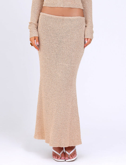 A person is wearing the CONNELL SKIRT - GOLD from TIGERMIST, a full-length textured skirt paired with a matching long-sleeve top made from knit fabric. Their feet in open-toe sandals are visible, and their midriff is slightly exposed. The background is a plain, light-colored studio setting.