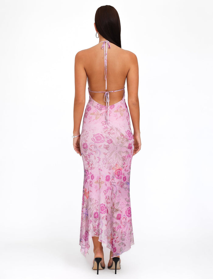 A woman stands facing away, showcasing the TIGERMIST COSETTE DRESS - PINK : DELICATE FLORAL with its versatile halter neckline and open back design. The flowing hemline adds an elegant touch to her look. She has long, dark hair and is wearing black high heels against a white background.