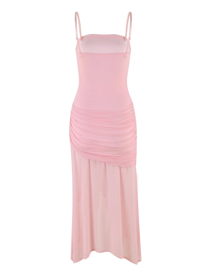 The COVE DRESS - PINK : BABY PINK from TIGERMIST is sleeveless with spaghetti straps, featuring a fitted silhouette and ruched detailing on the sides from the waist to the hips. It flows into a slightly flared, semi-transparent maxi skirt.