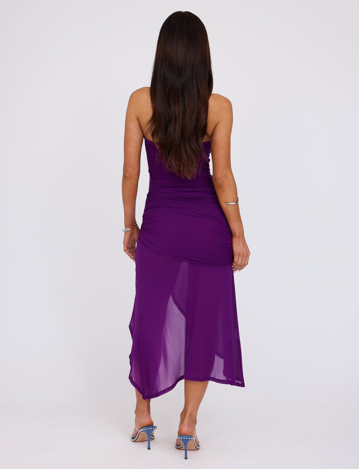 A woman with long, dark hair is standing, facing away from the camera. She is wearing a strapless COVE DRESS - PURPLE by TIGERMIST, featuring a ruched design with a sheer overlay and complemented by blue high-heeled sandals. The background is plain white.