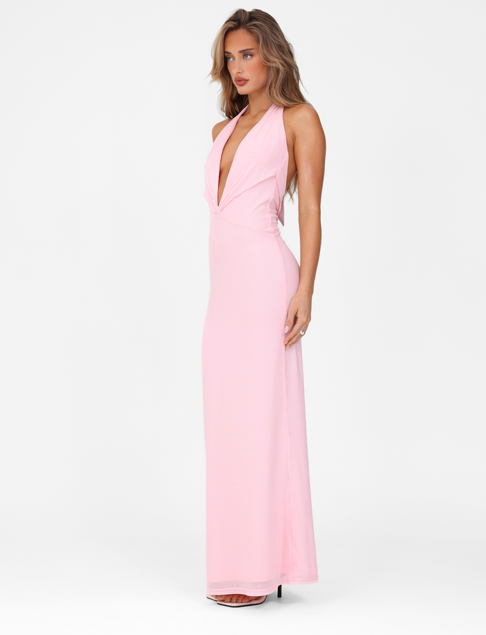 A woman stands against a white background, looking to the side in the TIGERMIST CRESSIDA MAXI DRESS - BABY PINK, a sleeveless halter neck dress with a deep V plunging neckline and maxi length.