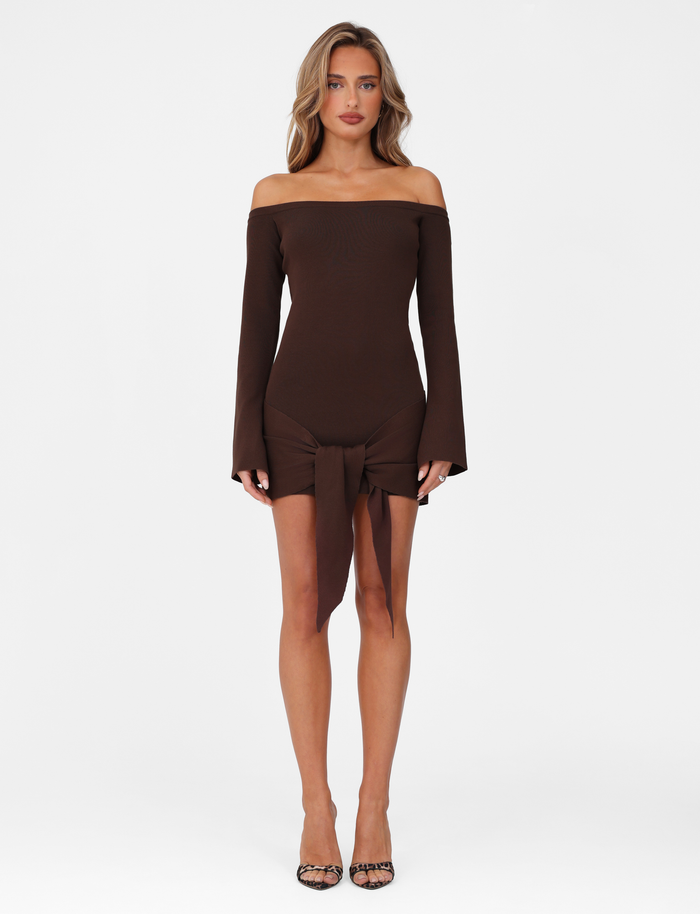 A woman poses against a white background in the TIGERMIST CYPRESS MINI DRESS - BROWN : CHOCOLATE, an off-the-shoulder, bodycon dress with a front tie detail. She pairs it with black open-toe heels and wears her hair loose and wavy.