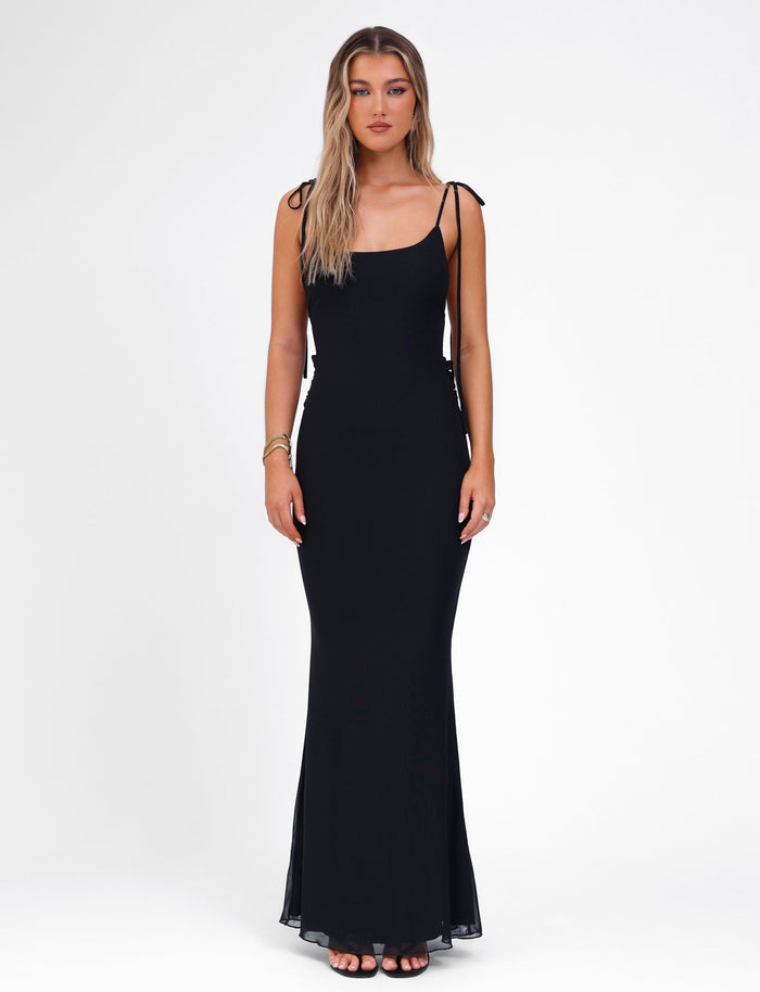 A woman with long blonde hair is wearing the CYRIL DRESS - BLACK by TIGERMIST. This sleeveless, form-fitting dress features adjustable straps and pairs seamlessly with her black high-heeled sandals. She stands against a plain white background, facing the camera with a neutral expression and minimal jewelry on her wrist and fingers.