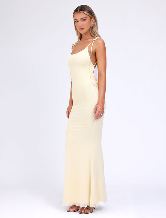A woman stands confidently against a plain white background, wearing the TIGERMIST CYRIL DRESS in lemon. The long, elegant maxi dress features adjustable straps, a side cut-out design, and a satin cowl back. She has long, straight hair and is looking directly at the camera with a calm expression.