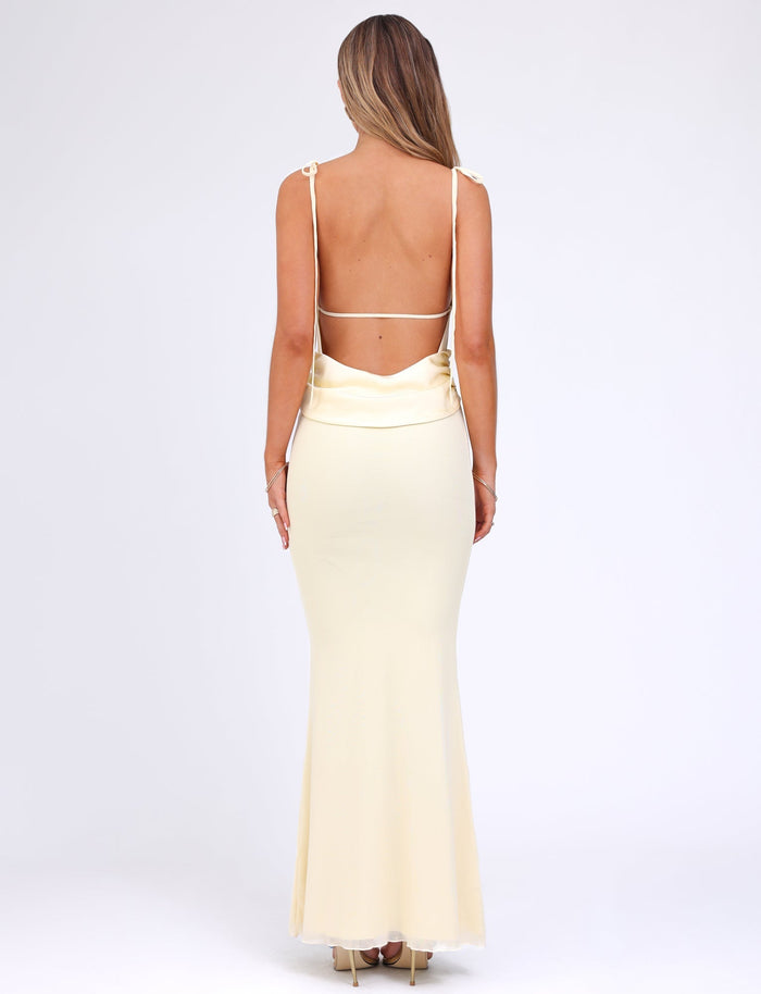 A woman with long blonde hair stands with her back to the camera, showcasing the CYRIL DRESS - YELLOW : LEMON by TIGERMIST, a stunning backless, floor-length lemon maxi dress featuring adjustable straps and a flowing hem. She is poised against a plain, light-colored background.
