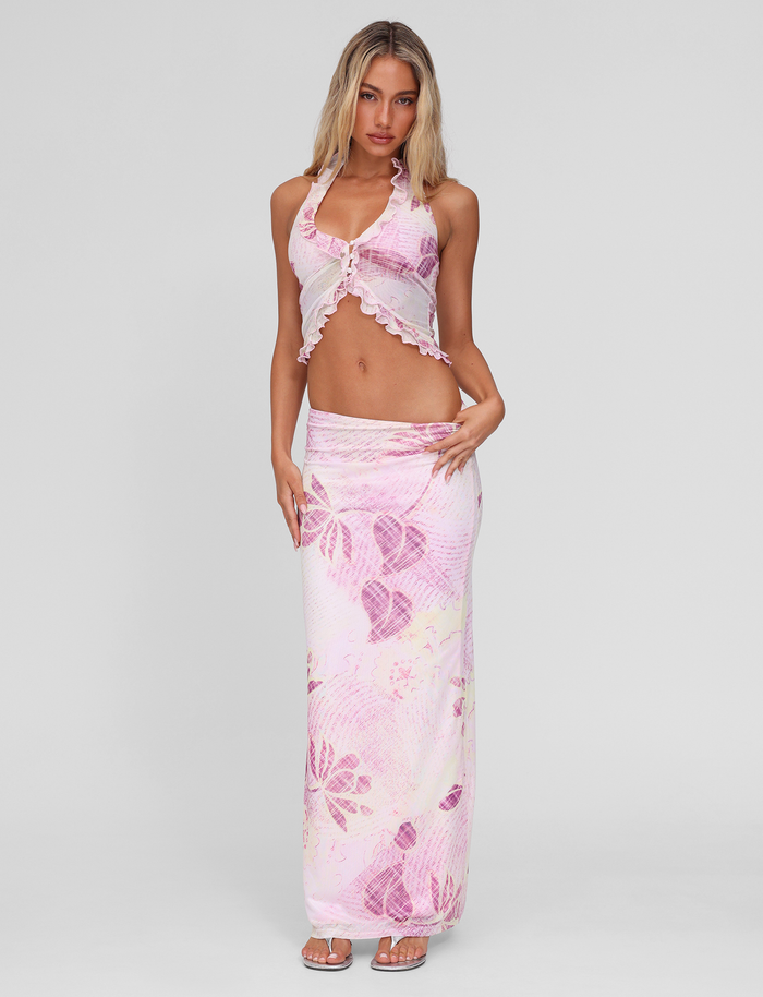 A woman stands against a plain background wearing the TIGERMIST DALILA MIDI SKIRT in Grunge Floral Magenta with a matching sleeveless halter crop top, both featuring ruffle trim, paired with sandals.