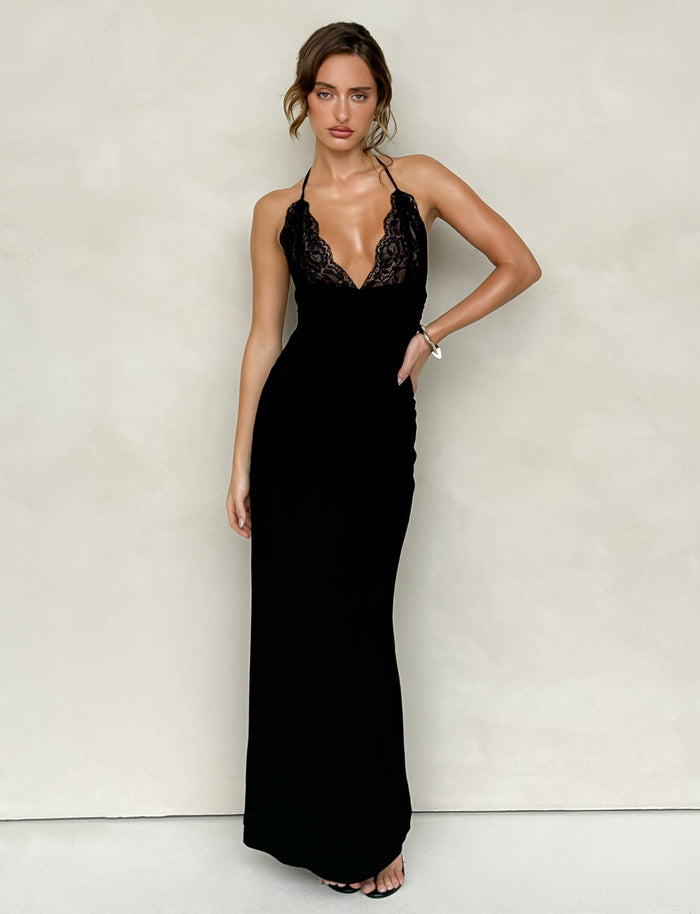 The individual is donning a gorgeous DEENA MAXI DRESS - BLACK by TIGERMIST, showcasing its long black design with intricate lace bust cups and a lace neckline. Posed against a neutral backdrop, their hair is elegantly styled up as they strike a pose with one hand on their hip.
