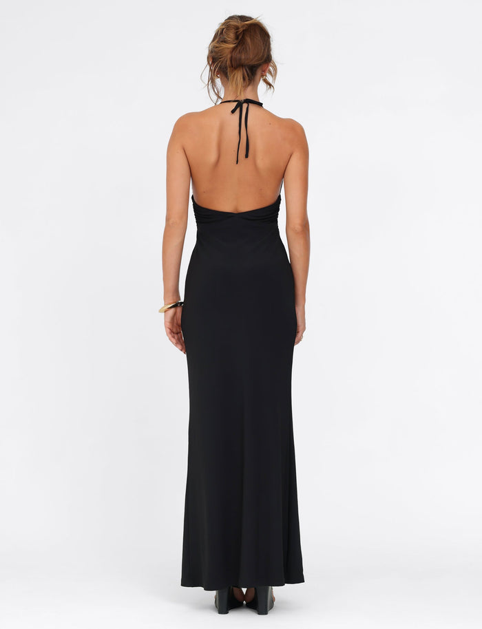 The scene captures a rear view of a person adorned in the elegant DEENA MAXI DRESS - BLACK by TIGERMIST, showcasing intricate lace bust cups. The long black backless halter neck design elegantly flows into a graceful maxi length. Their hair is styled in a loose updo as they stand poised against a plain white background.