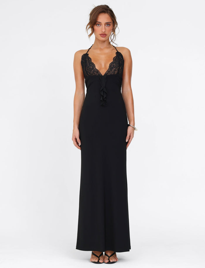 A woman stands against a white background wearing the DEENA MAXI DRESS by TIGERMIST. This elegant black dress features lace bust cups that beautifully enhance the neckline. Her hair is tied up, and she completes her look with black shoes and a silver bracelet, exuding timeless sophistication.
