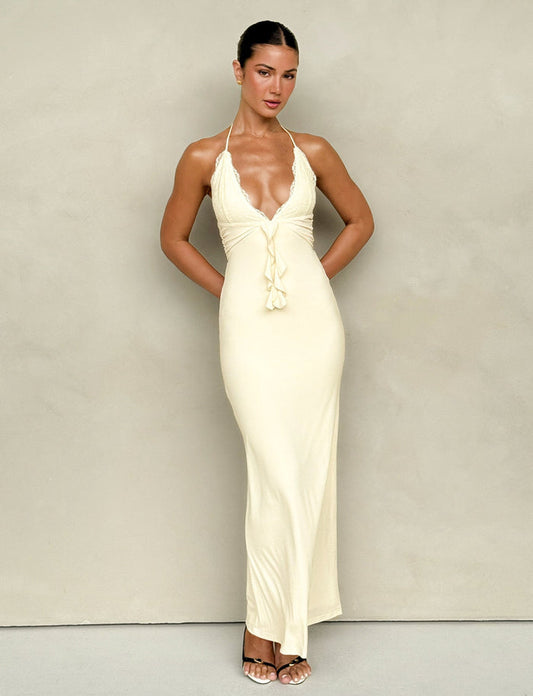 A woman poses against a solid backdrop, elegantly dressed in the DEENA MAXI DRESS - YELLOW : LEMON by TIGERMIST. This form-fitting halter dress, in a soft hue, features intricate lace bust cups and delicate ruffle details. The maxi-length attire is perfectly paired with her high-heeled sandals as she gracefully holds her hands behind her back.