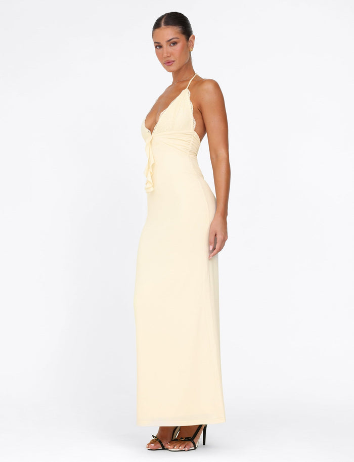 A woman poses against a white background in the sophisticated DEENA MAXI DRESS from TIGERMIST—a lemon yellow, backless halter dress featuring lace bust cups and a deep V-neckline. Her hair is pulled back as she stands in black high-heeled sandals, showcasing a side profile of the graceful maxi-length gown.