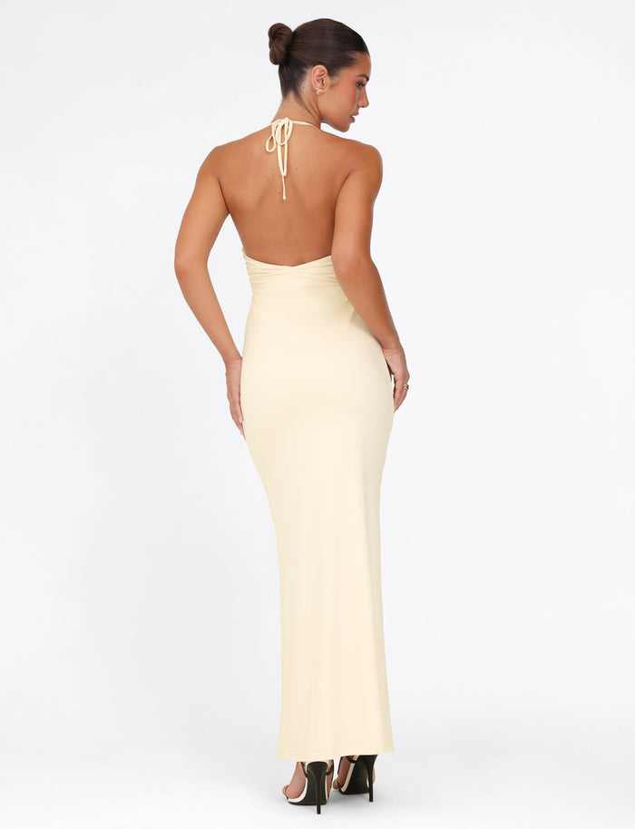 A woman in a sophisticated maxi-length DEENA MAXI DRESS - YELLOW : LEMON from TIGERMIST is turned away from the camera, showcasing its backless design and halter neck. Her hair is styled elegantly in an updo, and she complements the outfit with black high-heeled sandals. The lace bust cups enhance the dress's sophistication against the plain white background.