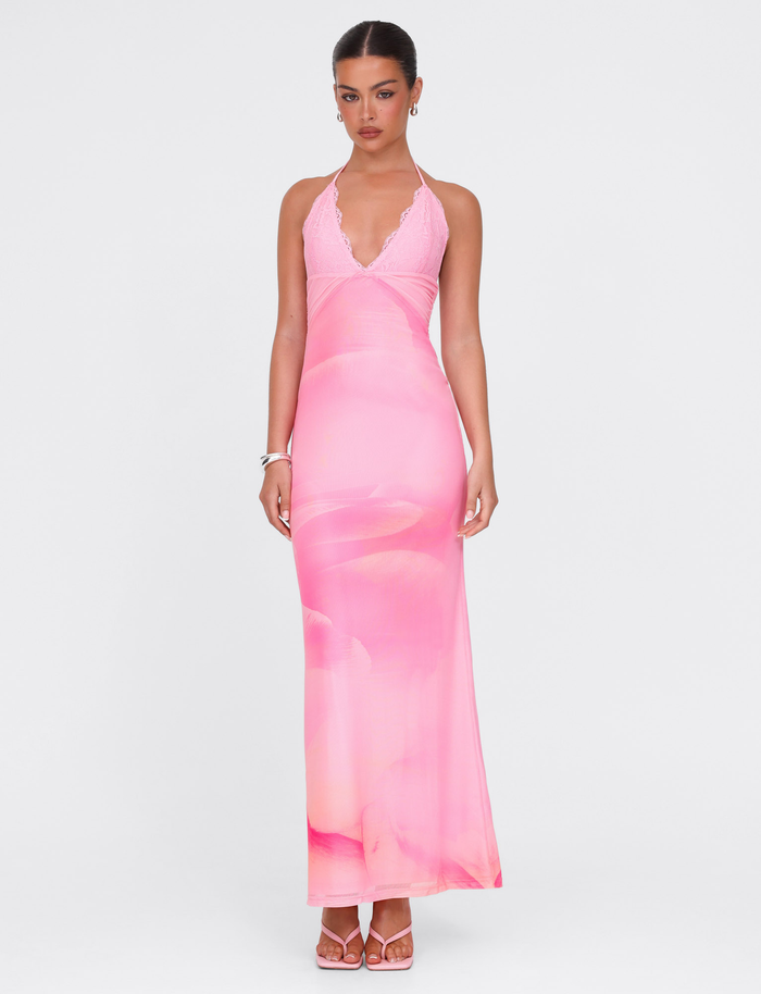 A woman stands against a white background wearing the TIGERMIST DENAE MAXI DRESS in Pink Blossom, featuring a floral design and delicate neckline. Her hair is tied back, and she accessorizes with pink sandals and a silver bracelet.