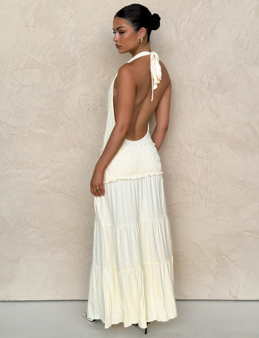 A woman stands sideways, looking back over her shoulder in the TIGERMIST DENALI HALTER DRESS - NEUTRAL: BONE, a cream-colored maxi with a fitted bodice and tiered skirt. Her hair is styled in a sleek bun against a light textured wall.