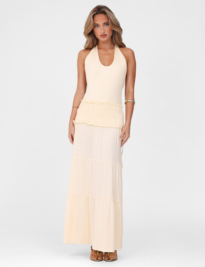 A woman with long wavy hair wears the TIGERMIST DENALI HALTER DRESS in Neutral: Bone, featuring a fitted silhouette and ruffled waist detail, standing against a plain white background and looking forward with a neutral expression.