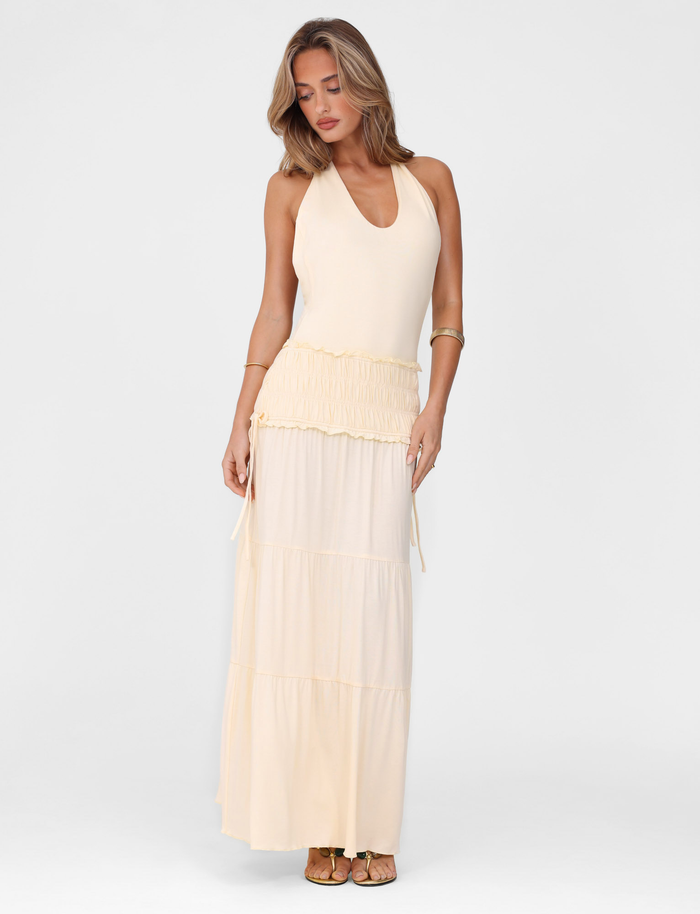 A woman models the TIGERMIST DENALI HALTER DRESS - NEUTRAL : BONE, a sleeveless maxi with a deep V-neck and tiered skirt, standing against a plain white background, looking down with one arm at her side and the other touching her waist.