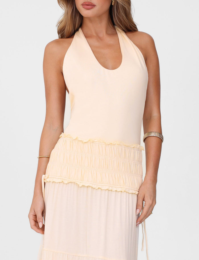 A woman wears the TIGERMIST DENALI HALTER DRESS in Neutral: Bone, a sleeveless maxi in buttery jersey with a gathered ruffled waist, fitted silhouette, and halter neckline. She accessorizes with bracelets against a plain white background.