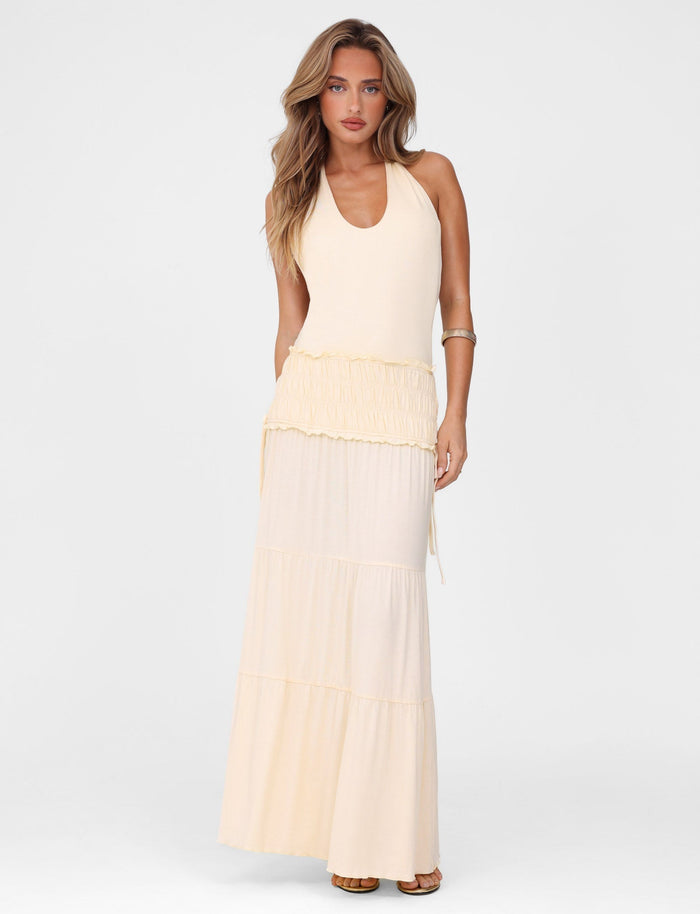 A woman with long wavy hair models the TIGERMIST DENALI HALTER DRESS in Neutral: Bone, a sleeveless maxi dress with a tiered full skirt, standing against a plain white background.