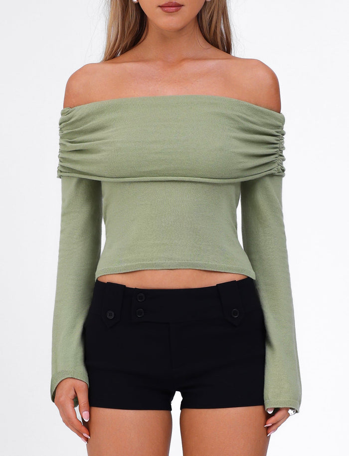 A woman is wearing the DIEDRE JUMPER - GREEN by TIGERMIST, an off-the-shoulder long-sleeve top made of high-stretch fabric, paired with black high-waisted shorts. The top features a gathered design at the shoulders, and her arms are relaxed at her sides. The background is plain white.