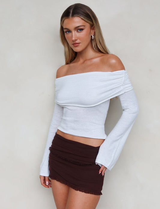 A woman with long blonde hair is standing against a neutral background. She’s wearing the DIEDRE JUMPER in white from TIGERMIST, featuring an off-the-shoulder neckline and long sleeves, paired with a short, high-stretch brown skirt. She has a stoic expression and is looking slightly off to the side.