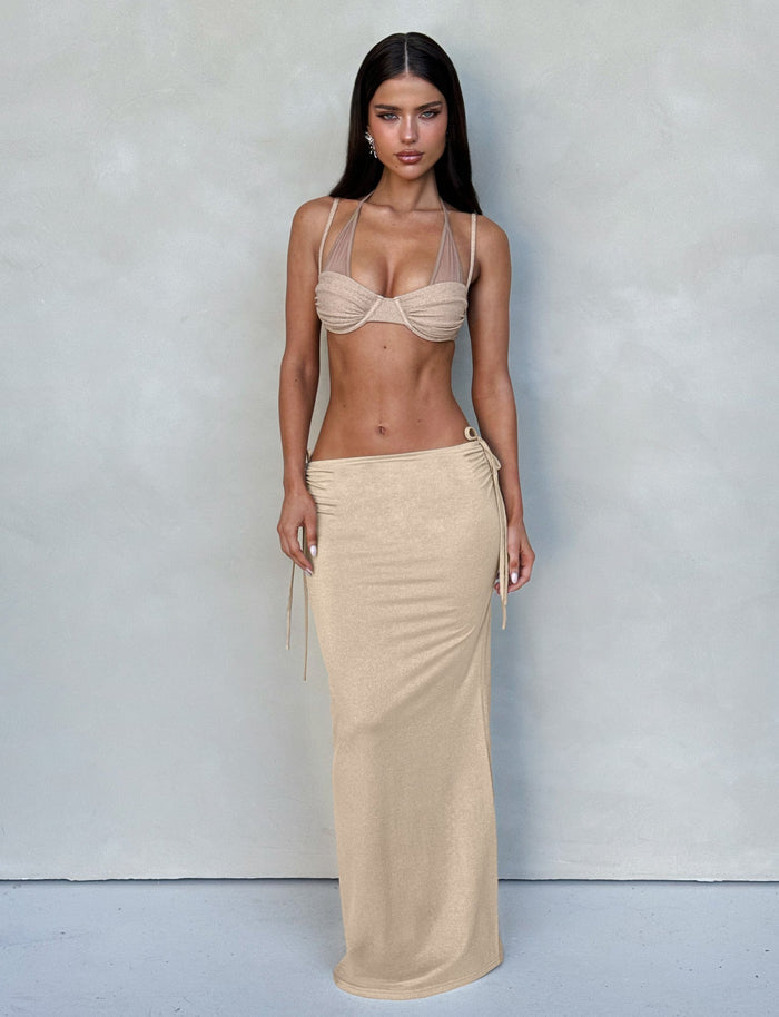 A woman with long dark hair is standing against a plain background. She's wearing the ELLIANA SKIRT - GOLD from TIGERMIST, paired with a matching beige bikini top featuring halter straps. She has a serious expression, and her hands are relaxed by her sides.