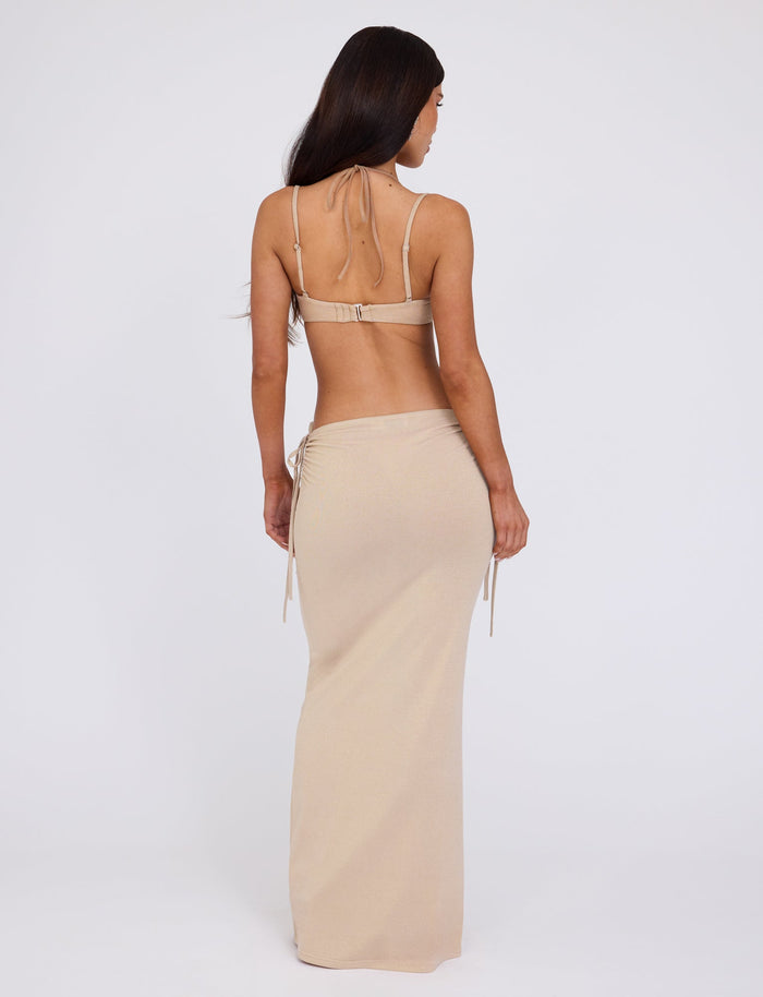 A woman with long, dark hair stands with her back to the camera against a plain white background, wearing a gold halter top that ties at the neck and the matching ELLIANA SKIRT from TIGERMIST.