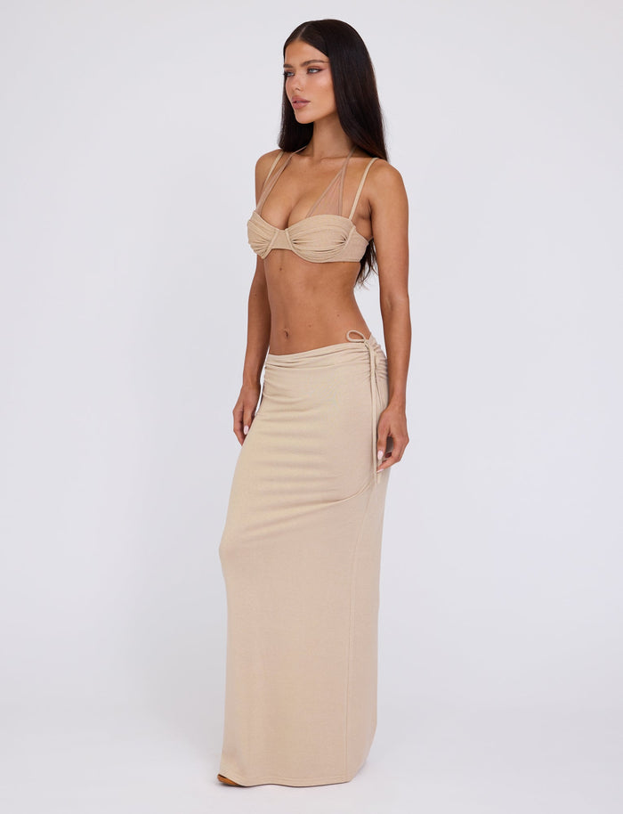 A woman stands against a neutral background wearing the TIGERMIST ELLIANA SKIRT in gold. The top is a strappy, bralette-style with ruched detailing, and the bottom is a long, fitted skirt with a side tie. She has long, straight, dark hair and a focused expression.