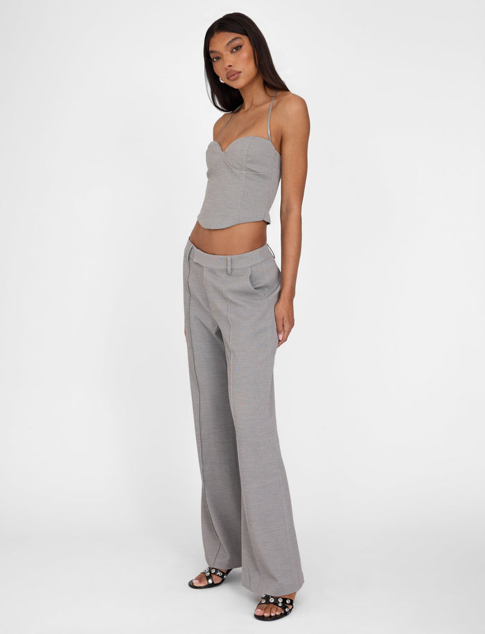 A woman poses in a light grey, spaghetti strap crop top with a sweetheart neckline and the EMALINE PANT - GREY by TIGERMIST. She stands with one arm bent at her waist and the other hanging by her side, wearing black heeled sandals, against a plain white background.