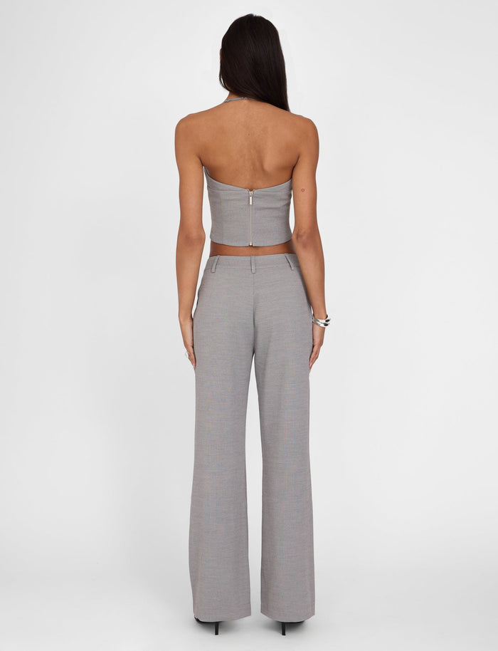 A woman with long dark hair is facing away from the camera. She is wearing a gray, strapless crop top with a back zipper and the EMALINE PANT - GREY from TIGERMIST. She accessorizes with silver bracelets and black high heels. The background is plain white.