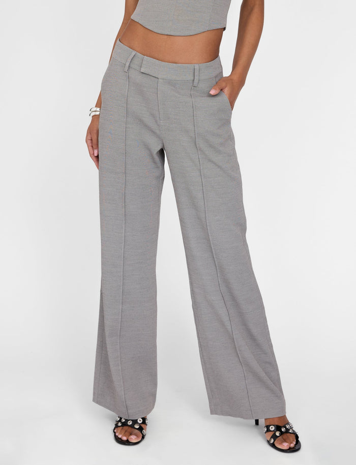 A person is modeling the TIGERMIST EMALINE PANT in grey with a tailored fit, their hands resting casually in their pockets. They are also wearing a matching grey crop top, accessorized with a bracelet on one wrist and black sandals featuring studded detailing. The background is plain white.