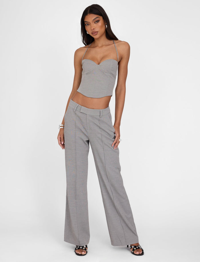 A woman poses wearing a grey two-piece outfit from TIGERMIST: a strappy crop top and the EMALINE PANT - GREY tailored-fit, wide-leg pants. She accessorizes with bracelets and black sandals. Her expression is neutral, and the background is a solid white.