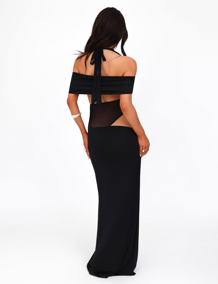 A woman with long dark hair is wearing the TIGERMIST EMBERLAIN DRESS - BLACK, featuring cut-out details at the waist and shoulder straps. She is facing away, showcasing the back of the maxi-length dress against a plain white backdrop.
