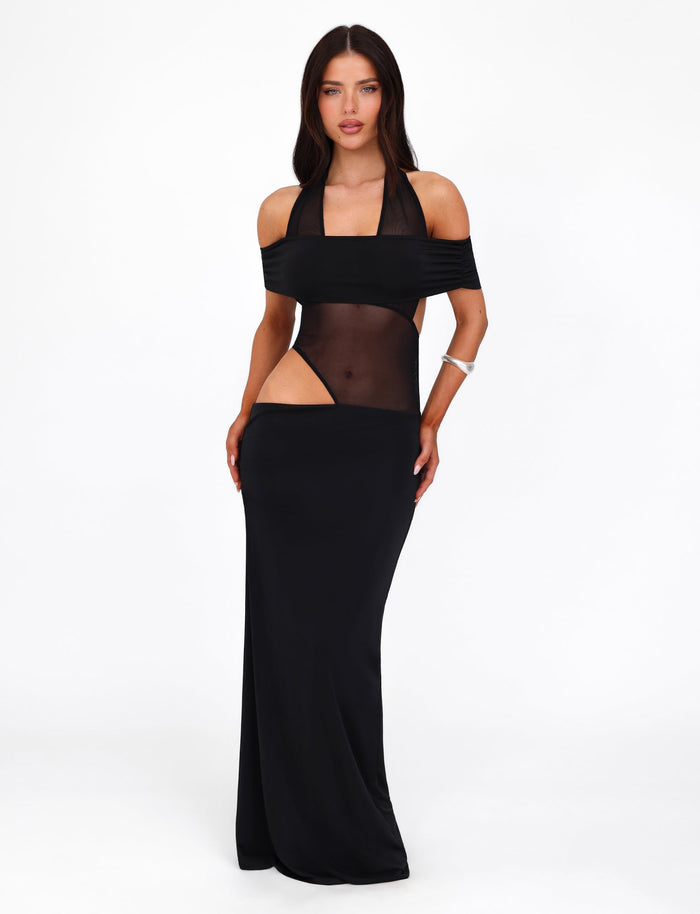 Against a white backdrop stands a woman donning the EMBERLAIN DRESS by TIGERMIST. The black dress features a halter neckline and a mesh panel that offers an alluring glimpse of skin. Her long, dark hair flows past her shoulders, perfectly complementing the dress's maxi length. She accessorizes with a silver bracelet on her right wrist and maintains a neutral expression as her hands rest on her thighs.