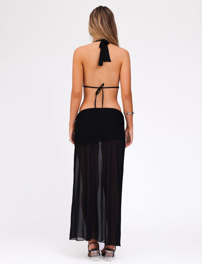 A woman with long hair is standing with her back to the camera. She is wearing the ESHA MAXI DRESS - BLACK by TIGERMIST, a stunning black halter neck dress that flows elegantly to maxi length with a sheer skirt. She has a bracelet on her left wrist and is standing against a plain white background.